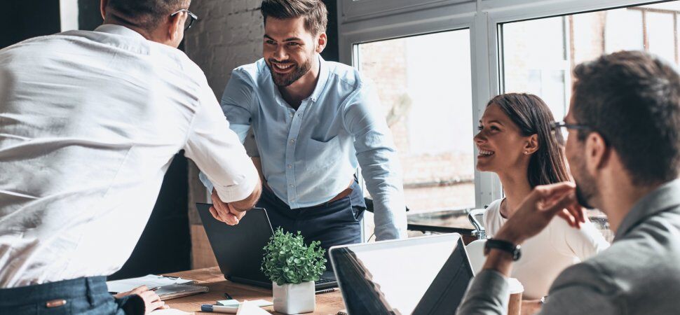 Businessman and businesswoman smile at two businessmen shaking hands at a meeting