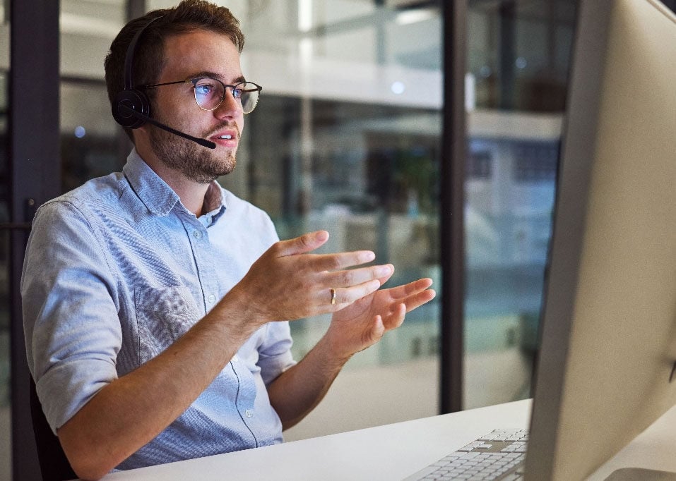 Man wearing glasses and a headset with a microphone gestures at a computer screen in his business office